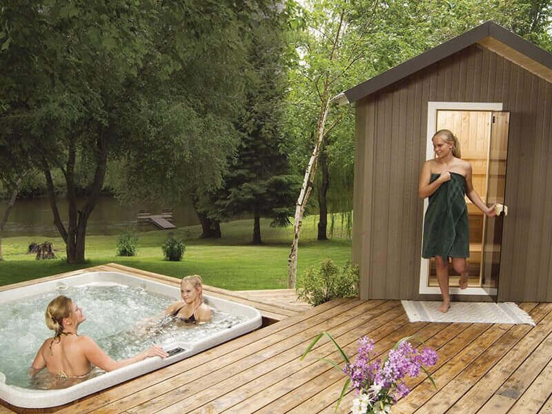 Woman exiting outdoor Finnleo sauna with towel wrapped around her while husband and child soak in a hot tub