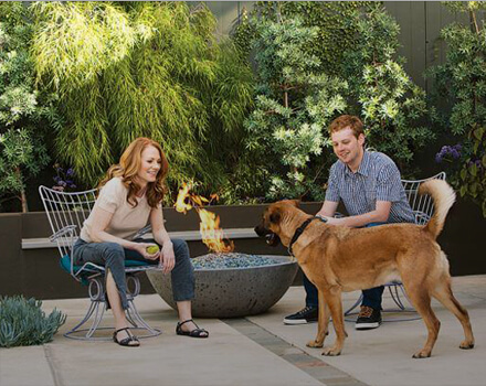 Man and woman petting dog in backyard next to firepit while sitting on outdoor furniture